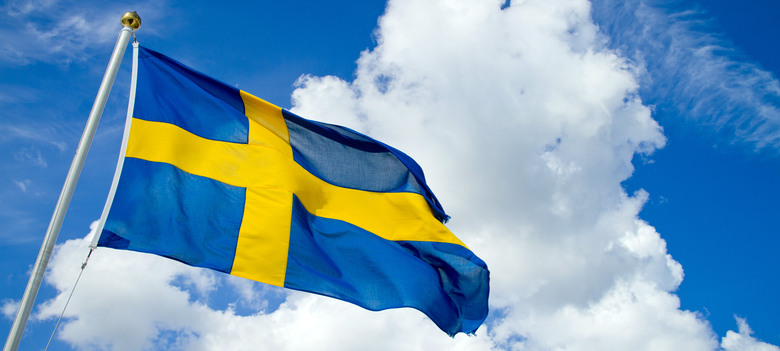 Svenska flaggan vajer i vinden. Bakom flaggan syns en blå himmel och vita moln.