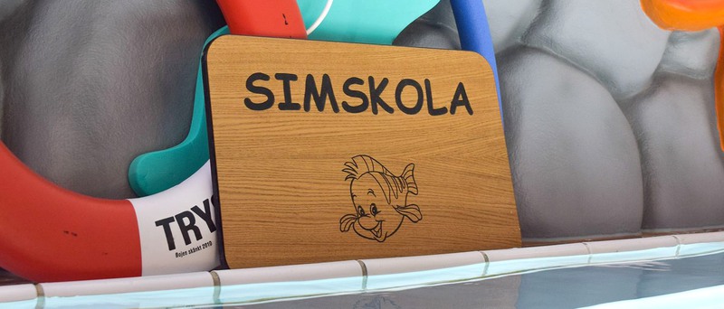 Skylt där det står simskola. Badflytsaker i bakgrunden.