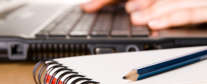 Block och penna och i bakgrunden syns händer som skriver på en dator.