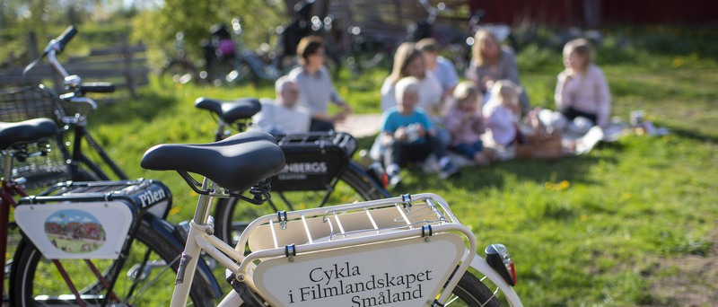 En cykel i förgrunden där det står "Cykla i Filmlandskapet Småland. I bakgrunden sitter några vuxna och barn på en filt i gräset.