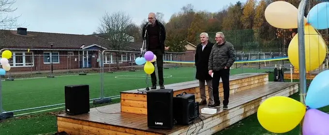 Rektor talar i mikrofon och två politiker står bakom på skolgård.